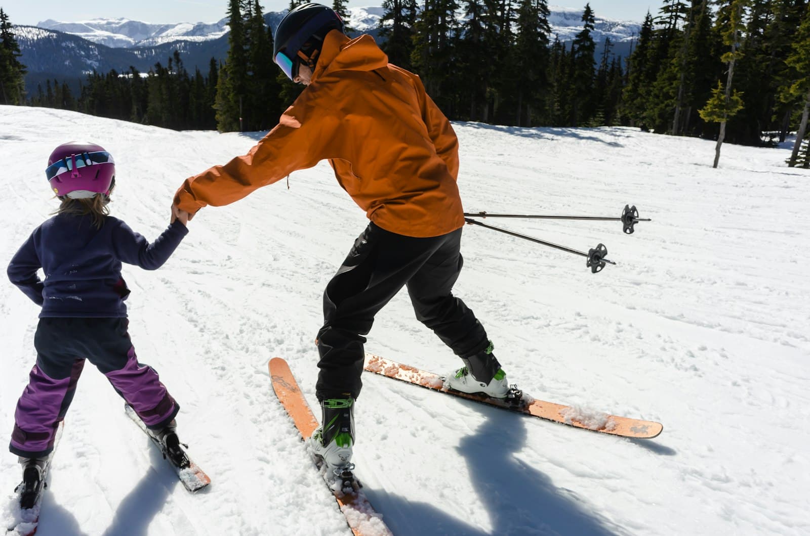 Volwassene in oranje jas houdt de hand vast van een klein kind op ski's